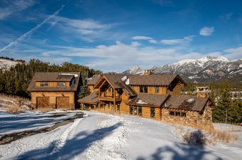 The Latigo Lodge, a grand wooden mountain home, stands majestically in a snow-covered landscape with towering pine trees and snow-capped mountains under a clear blue sky.