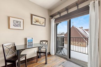 The dining area in Cana Loft 203 features a dark wood table and chairs, with framed art on the wall and a sliding glass door opening to a snow-covered balcony view of buildings and a rooftop cross.
