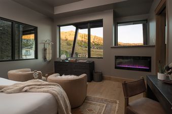 A serene bedroom at Aria Ridge with a cozy bed, two plush chairs, a modern electric fireplace, and large windows offering a breathtaking mountain view.