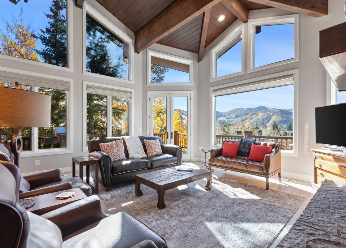 The living area at Bear Hollow 5428 features an impressive vaulted ceiling with exposed wooden beams, large windows offering panoramic views of the surrounding mountains and autumn foliage, and a comfortable seating arrangement with leather sofas and a ru