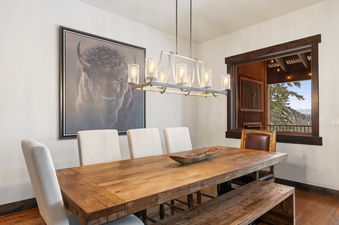 The dining area at Inspiration Ski Haus features a rustic wooden table, upholstered chairs, a matching bench, a linear chandelier, and a framed bison painting on the wall, with a window offering a view of a snowy landscape.