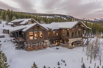 A luxurious, multi-story wooden chalet with snow-covered roofs is nestled among pine trees in a vast, snowy landscape under a soft, cloudy sky.