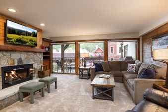 A cozy living room at Montaneros 317 featuring an L-shaped sectional sofa, a stone fireplace with a fire, and large windows looking out onto a green landscape and balcony.