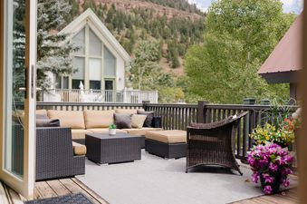 An outdoor patio at Smugglers Hideaway with wicker furniture, beige cushions, and potted flowers, overlooking a treed landscape and a distant mountain.