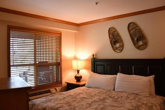 A cozy bedroom with a large bed, a nightstand with a lamp, a window with blinds, and decorative snowshoes on the wall.