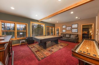 A spacious game room at the Baldy Ridge Chalet, featuring a pool table, shuffleboard, and comfortable seating, with large windows overlooking a snowy mountain landscape.