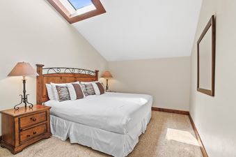 A cozy bedroom at Fairway Village 2919 featuring a bed with white linens, wooden nightstands with lamps, a skylight, and a mirror on the wall.