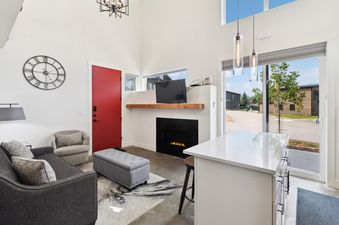A modern and bright living area featuring a grey sofa, a fireplace with a TV above, and large windows with outdoor views.