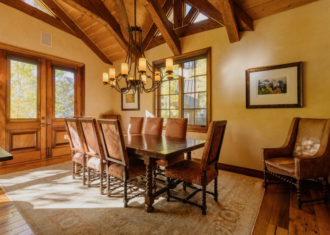 A rustic dining room with a large wooden table, upholstered chairs, a chandelier, and windows looking out onto autumn trees.