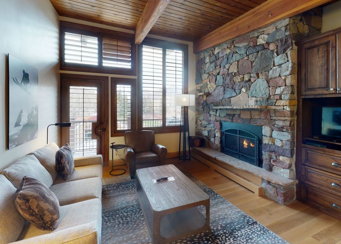 Relax in the living room of Manor Vail 360, which features a fireplace, a large sectional sofa, a coffee table, a leather armchair, and large windows offering views of the surrounding mountains.