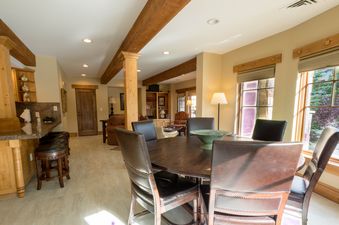 A spacious dining and living area in The Chateaux Three Bedroom Suite features a wooden dining table, leather chairs, and large windows with an exterior view.