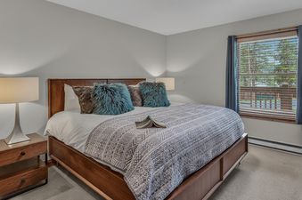A cozy bedroom at First Tracks Chalet with a wooden bed, patterned gray duvet, turquoise pillows, and a window showing pine trees.