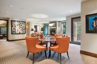 A spacious living area within The Winter Watch Estate featuring orange leather chairs, a round wooden table, a stone fireplace with a television, and large windows revealing an outdoor view.