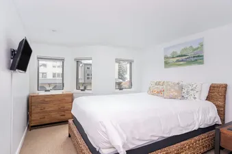 The bright bedroom of Lulu Luxe features a comfortable bed with decorative pillows, a wall-mounted TV, a wooden dresser, and two bay windows offering an exterior view.