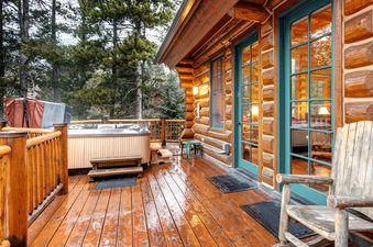 A rustic wooden deck features a hot tub with an open cover, surrounded by pine trees, and teal-framed glass doors leading into Obsidian Jackson Hole.