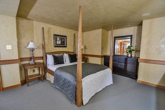 A four-poster bed with white bedding and dark accent pillows is centered in a cozy bedroom with golden textured walls and wooden trim, flanked by nightstands with lamps.