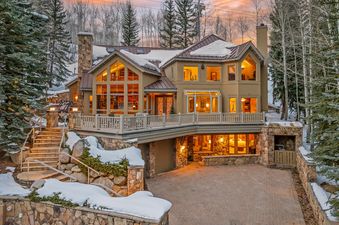 Founders Lodge is a luxurious mountain home with a snow-covered roof and yard, stone steps, and a large deck with a view.