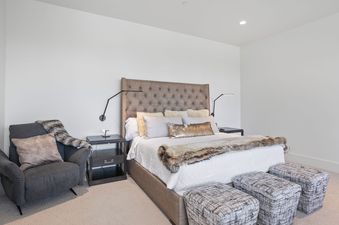 A primary bedroom at Quicksilver Haus features a grand bed with a tufted headboard, flanked by modern task lamps and dark wood nightstands, and includes a comfortable armchair and patterned ottomans.
