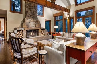 The living room of A Creekside Lodge features a large stone fireplace with a roaring fire, comfortable seating, and large windows offering a scenic view.