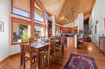 A spacious dining area with a wooden table and chairs is connected to a kitchen, featuring large windows with wooden frames and a high wooden ceiling, creating an inviting open-concept living space in Crescent Cabin.