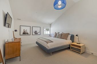 A serene guest bedroom in Stillson Meadow features a plush bed with blue frame, soft white bedding, and earth-toned pillows, complemented by wooden nightstands, modern lamps, and animal-themed framed art on white walls, with a blue pendant light and a wal