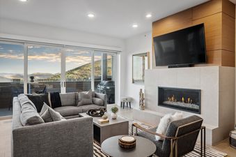 The luxurious living room of Apex 208 features a large gray sectional sofa, a modern fireplace with a TV mounted above, and floor-to-ceiling windows offering stunning mountain views.