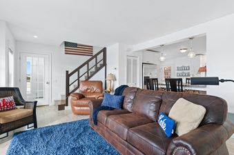 An inviting living room at the Lake Blaine Lake House, featuring a brown leather sofa, an armchair, a blue rug, and an American flag decor, with an open-concept kitchen visible in the background.