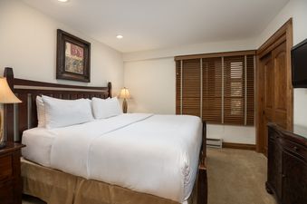 A cozy bedroom at Montaneros 321 featuring a large bed with white linens, a dark wood headboard, bedside lamp, and wooden blinds.