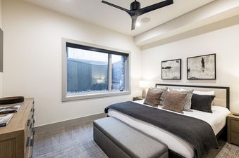 A modern bedroom at Frostwood Links with a large bed, decorative pillows, a sleek ceiling fan, and a unique window view of a rock-filled outdoor area.