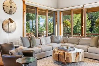 A cozy living area at Telemark A with a large sectional sofa, a unique live-edge coffee table, and large windows overlooking a lush green exterior with hanging flower baskets.