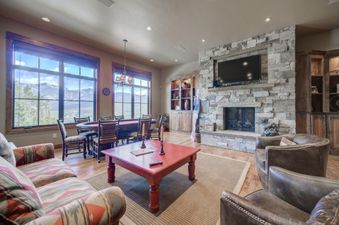 The grand living area of Peaks View Lodge features large windows showcasing mountain views, a stone fireplace, comfortable seating, and a gaming table.