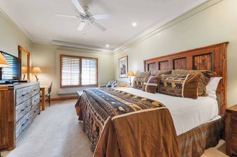 The master bedroom features a king-size bed with a rustic wooden headboard, a large dresser with a TV, and a desk, all illuminated by natural light from a window with wooden blinds and ambient lighting.