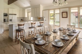 A bright, open-concept kitchen and dining area at Glenn House, featuring white cabinetry, a large kitchen island with bar stools, and a dining table set for many guests with large windows overlooking the outdoors.