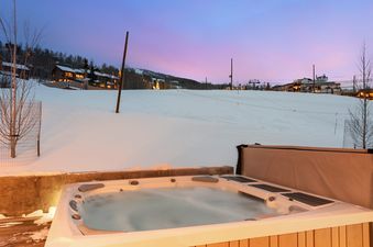 A steamy hot tub on a wooden deck with a snow-covered mountain and illuminated buildings in the background under a purple and blue sunset sky.