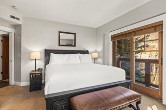 A spacious bedroom at Town Pointe C101 with a large bed, nightstands, lamps, a leather bench, and a sliding glass door leading to a balcony.