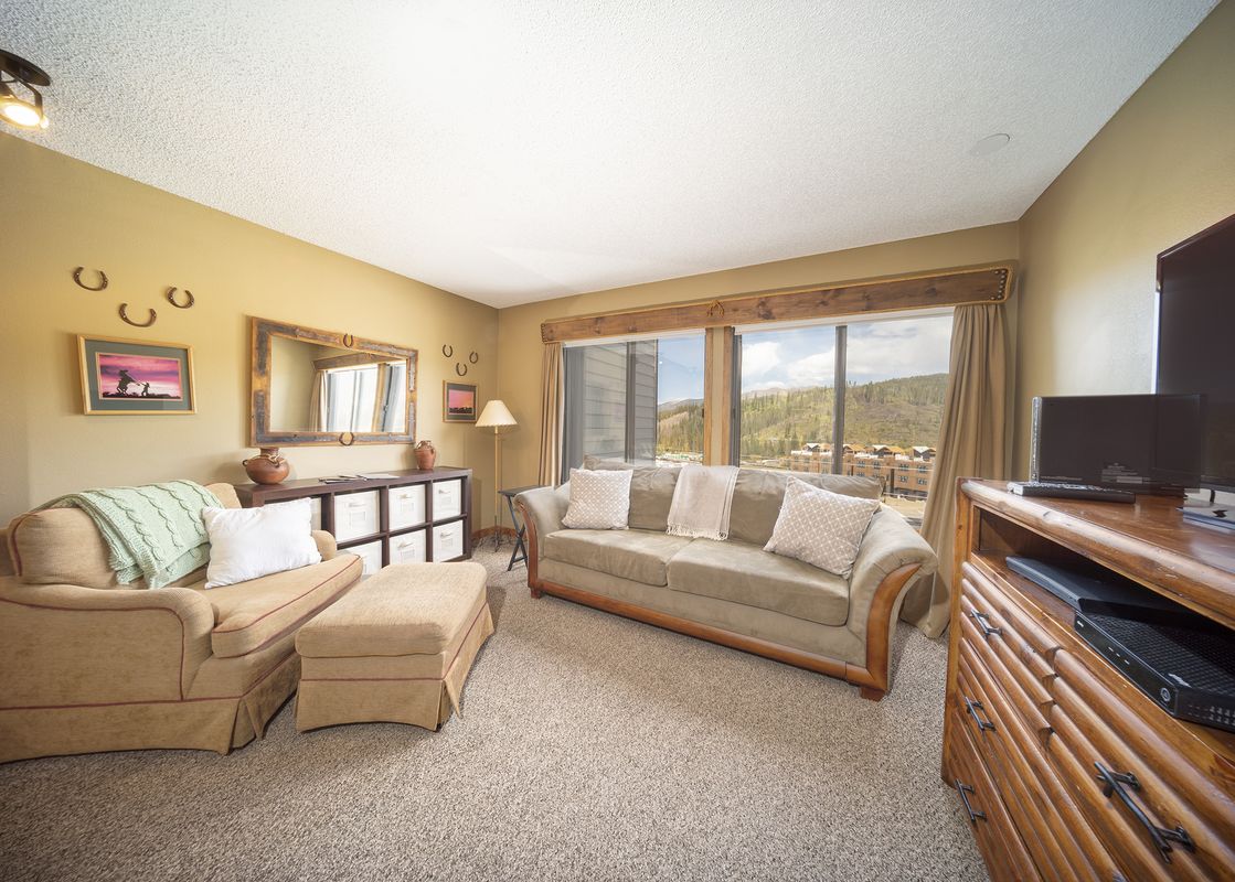 The living area features a comfortable armchair, a sofa with throw pillows, and a wooden entertainment stand with a television, all set on a light brown carpet.