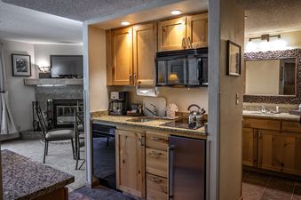 A compact kitchenette with wooden cabinets, a microwave, a dishwasher, and a refrigerator, adjoined by a living area with a fireplace and a bathroom with a mirrored vanity.