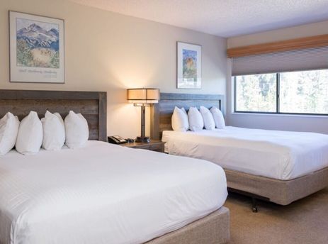 A hotel room at Beaver Run Resort with two beds, each featuring a rustic wooden headboard and pristine white bedding, alongside a bedside table with a lamp and telephone, and a window offering a view of lush trees.