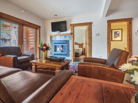 A cozy living room in the Mountain Lodge One Bedroom Deluxe Condo with brown leather furniture, a stone fireplace, and a flat-screen TV, offering a warm and inviting atmosphere.