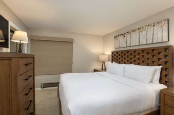 A king-size bed with white bedding and pillows, flanked by a wooden nightstand and lamp, with a stylish wooden headboard and a birch tree art piece above.