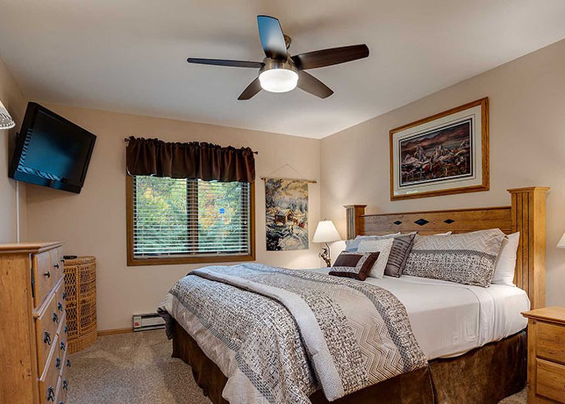 A serene bedroom at Ranch at Steamboat RA401 features a comfortable bed, rustic wooden furniture, a ceiling fan, and a wall-mounted TV, with a window offering a view of the outdoors.