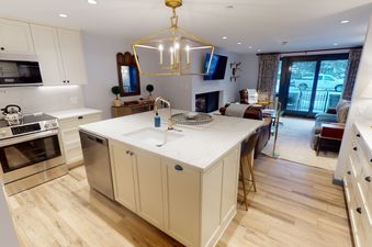 A kitchen with stainless steel appliances and white cabinets. There is a large island in the center of the kitchen with a white countertop and a sink. The kitchen opens up to a living room with a fireplace, a couch and a view of the outside.