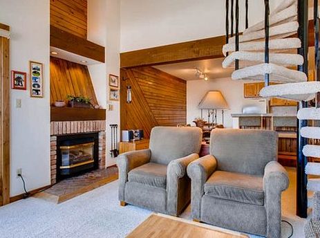 A cozy living room at Buttes 2 Peak 517 featuring a brick fireplace, two armchairs, a spiral staircase, and a glass door leading to an outdoor balcony with tree views.