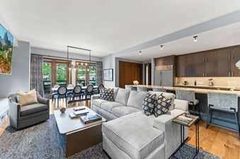 A spacious living room with a white sectional sofa, a coffee table, a large window with a view of the mountains, and a dining table in the background.