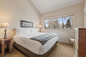 A serene bedroom in Snow Flower 127 featuring a queen bed with white linens, a grey throw, a rustic nightstand with a ski-themed lamp, and a large window overlooking snow-covered trees.
