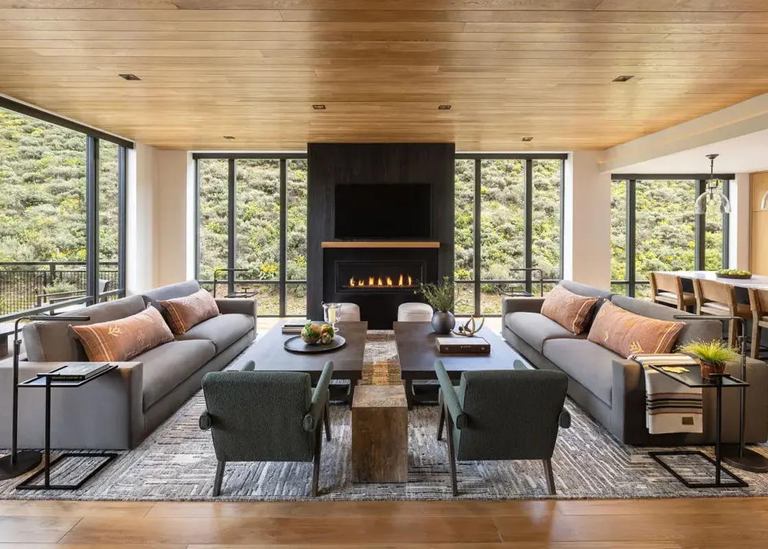 A wide view of the modern and spacious living room in The Sylvan Lodge Four Bedroom Junior Penthouse, featuring gray sofas, green armchairs, a gas fireplace with a TV above, and large windows with scenic mountain views.