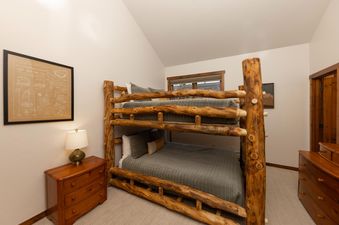 A rustic bunk bed with a wooden frame is featured in a cozy bedroom, complete with soft gray bedding, a bedside table with a lamp, and a framed art piece on the wall.