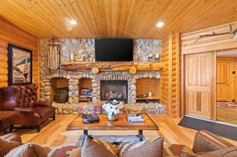 The image showcases the cozy living room of Black Bear 308, featuring a stone fireplace with a wooden mantel, a flat-screen TV, comfortable leather seating, and rustic wooden decor, creating a warm cabin atmosphere.