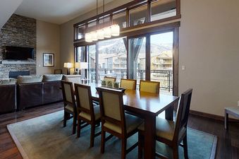 The dining area at Trailhead Lodge 4100 features a dark wood table and chairs with light cushions, set against large windows with views of other buildings, and illuminated by natural light and contemporary lamps, with a stone fireplace at one end.