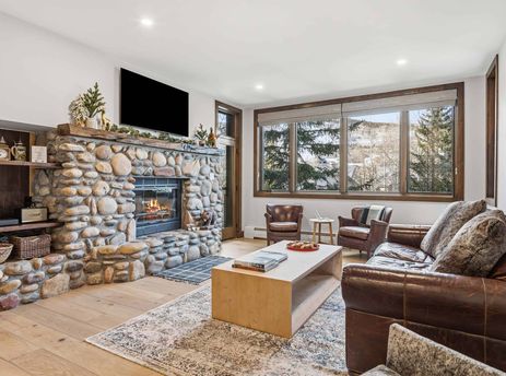 A cozy living room at Highlands Lodge 205 features a stone fireplace, leather furniture, and large windows with a view of snow-dusted evergreen trees and mountains.